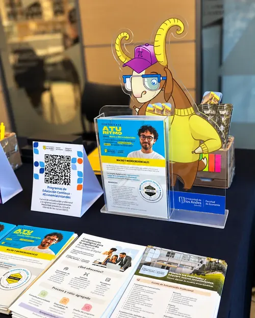 Exhibidor acrílico con la mascota de la Facultad de Economía en suéter amarillo y gorra, junto a folletos de microcredenciales de Educación Continua